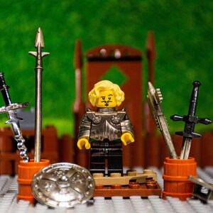 Puede incluir: Una minifigura Lego amarilla con armadura plateada y una capa negra se encuentra frente a un trono de madera. La minifigura sostiene una espada y un escudo. Hay otras dos espadas en el fondo.