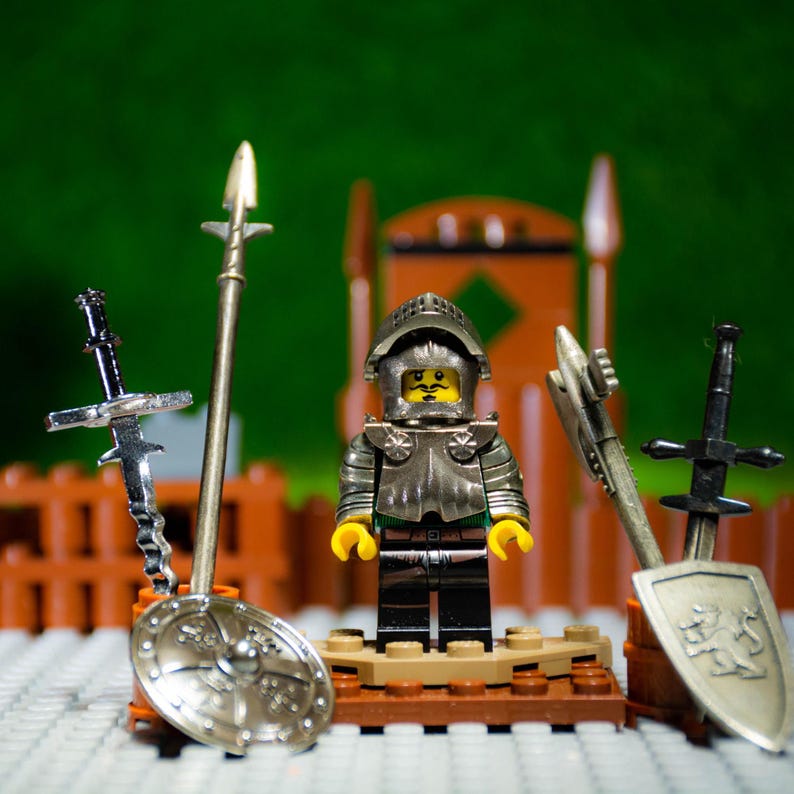 Puede incluir: Una figura de caballero Lego con armadura plateada, cara amarilla y capa verde, se encuentra sobre una base marr&oacute;n. El caballero sostiene una espada plateada y est&aacute; rodeado de otras armas plateadas, incluyendo una lanza y un escudo.