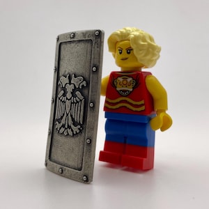 May include: A silver shield with a black and white eagle design. The shield is held by a yellow Lego minifigure with blonde hair, wearing a red shirt with a yellow and orange design, blue pants, and red shoes.