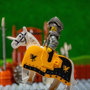 Puede incluir: Un caballero Lego con armadura plateada montando un caballo blanco con una silla de montar amarilla y negra. El caballo tiene un dise&ntilde;o de p&aacute;jaro negro en la silla de montar.