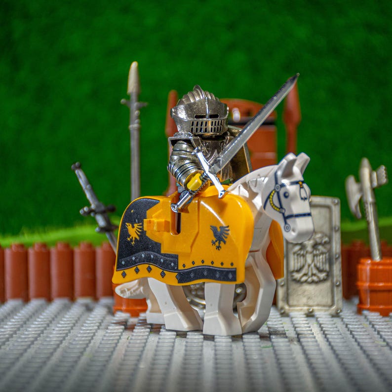 Puede incluir: Una figura de caballero de plata montando un caballo blanco con una silla de montar amarilla y negra. El caballero sostiene una espada de plata y lleva un casco de plata con una visera negra. La figura est&aacute; sobre una base de Lego gris y blanca.