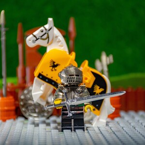 Puede incluir: Una figura de caballero plateado sosteniendo una espada plateada, de pie frente a una figura de caballo blanco y amarillo. El caballo tiene un dise&ntilde;o de p&aacute;jaro negro en su pecho.