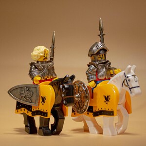 Puede incluir: Dos minifiguras Lego vestidas como caballeros a caballo. Los caballeros llevan armadura plateada y empu&ntilde;an espadas. Los caballos son negros y blancos y tienen mantas de silla amarillas y negras. Los caballeros miran hacia adelante.