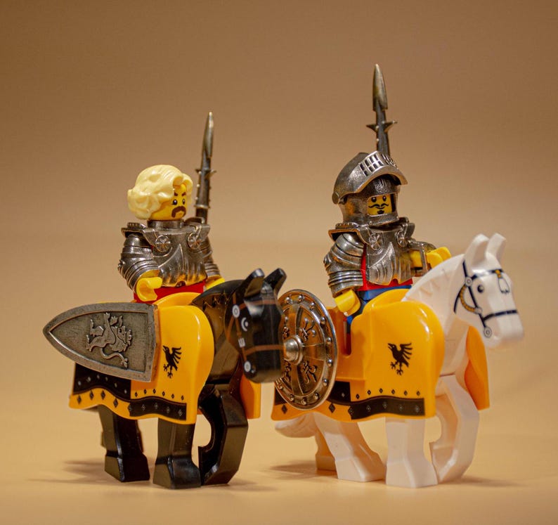 Puede incluir: Dos minifiguras Lego vestidas como caballeros a caballo. Los caballeros llevan armadura plateada y empu&ntilde;an espadas. Los caballos son negros y blancos y tienen mantas de silla amarillas y negras. Los caballeros miran hacia adelante.