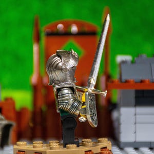Puede incluir: Una figura de caballero de juguete de plata con una espada y un escudo, de pie sobre una base marr&oacute;n. El caballero lleva un casco y una armadura. La figura est&aacute; delante de un fondo verde.