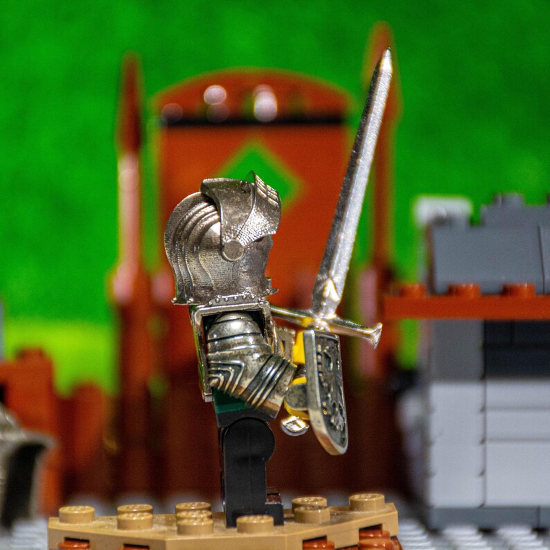Puede incluir: Una figura de caballero de juguete de plata con una espada y un escudo, de pie sobre una base marr&oacute;n. El caballero lleva un casco y una armadura. La figura est&aacute; delante de un fondo verde.