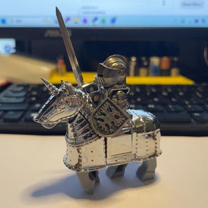 May include: A silver-colored knight riding a unicorn toy. The knight is wearing a helmet and armor, holding a sword and shield. The shield has a decorative emblem. The unicorn has a horn and is also silver.