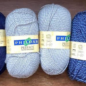 Pode incluir: Seis novelos de lã em tons de azul e cinza. A etiqueta indica "PHILDAR PREFACE". Cada novelo pesa aproximadamente 50 gramas. A lã está bem enrolada, ideal para projetos de tricô ou crochê. Estão dispostos sobre uma superfície de madeira.