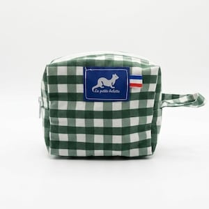 Peut inclure: Une petite trousse de toilette carrée à carreaux verts et blancs. Un écusson bleu présente une illustration de furet blanc et le texte "La petite belette". Une petite poignée et une fermeture éclair sont visibles.