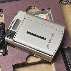 May include: A silver Olympus Pearlcorder L400 microcassette recorder featuring a small digital display, a black speaker grille, and control buttons labeled REC, STOP, PLAY/DIR, and FF/REW. A black wired accessory is attached to the side of the device.