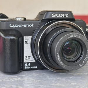 May include: A black Sony Cyber-shot digital camera featuring a 10x optical zoom lens, a 3.0 inch LCD monitor, and 8.1 megapixels. The lens is labeled Carl Zeiss Vario-Tessar 3,5-4,4/6,3-63. A black camera strap is attached to the side of the device.