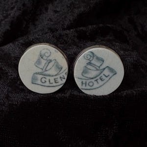 May include: Two round, white cufflinks with a silver border. Each cufflink features a blue illustration of a banner with the text "GLEN HOTEL". The background is a dark, textured fabric.