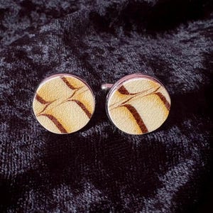 May include: A pair of round cufflinks with a cream-colored, textured surface and dark brown, curved vein-like patterns. The cufflinks have a silver-colored metal border and post.