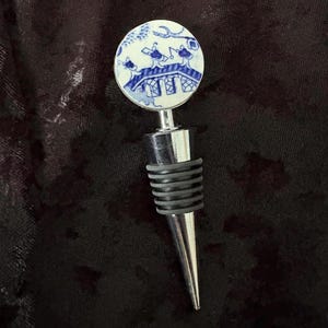 Może przedstawiać: Dekoracyjny korek do butelki z niebiesko-białą porcelanową górą z tradycyjnym wzorem inspirowanym Azją. Korek ma srebrny metalowy trzpień, czarną gumową uszczelkę i srebrną stożkową końcówkę.