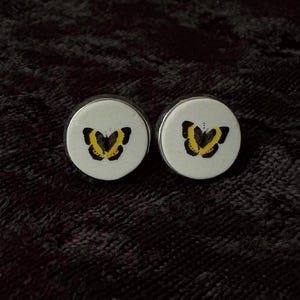 May include: A pair of circular stud earrings featuring a white face with a black and yellow butterfly graphic centered on each piece. The earrings have a smooth, rounded profile and a clean, high-contrast design on the front surface.