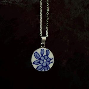 Puede incluir: Un collar de cadena de metal plateado con un colgante circular. El colgante tiene una superficie blanca similar a la cerámica decorada con un patrón floral azul pintado a mano, que consiste en una flor central rodeada de varios pétalos alargados.