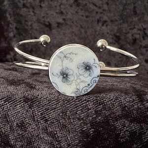 May include: Silver cuff bracelet with a circular floral design. The central disc has a light background with a black and grey floral pattern. The bracelet has a double band and rounded terminals.