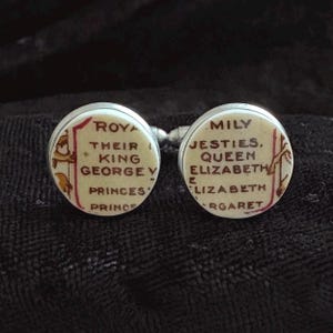 Może przedstawiać: Para okrągłych spinek do mankietów o kremowej powierzchni z brązowym nadrukiem. Lewa spinka zawiera słowa ROYAL, THEIR, KING, GEORGE V, PRINCES, PRINCE. Prawa spinka zawiera słowa MILY, JESTIES, QUEEN, ELIZABETH, ELIZABETH, RGARET.