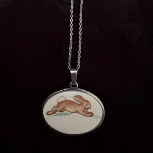 May include: A silver necklace with an oval pendant. The pendant depicts a brown rabbit running across a patch of green grass. The pendant has a white background and a silver border. The chain is delicate.