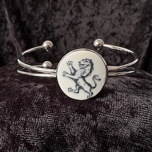 May include: Silver-toned cuff bracelet with a circular, off-white centerpiece. The centerpiece features a black illustration of a lion. The bracelet has a double-band design with small, spherical accents at the ends.