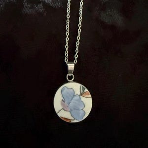 Könnte beinhalten: Eine silberfarbene Halskette mit einem runden Anhänger. Der Anhänger ist weiß und mit einer handgemalten hellblauen Blume sowie dezenten braunen Blattdetails verziert. Eine Metallöse verbindet das runde, keramikartige Schmuckstück mit der Kette.