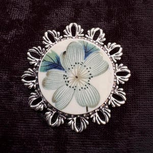 May include: A circular brooch featuring a central white disc with a printed light blue and green flower illustration. The disc is encased in a decorative silver-toned metal frame with an intricate, scalloped, and openwork filigree border design.