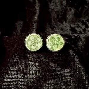 May include: A pair of circular stud earrings featuring a light green surface with a dark green botanical pattern of four-petaled flowers and dotted textures, encased in a thin metallic rim with a polished silver-toned finish.