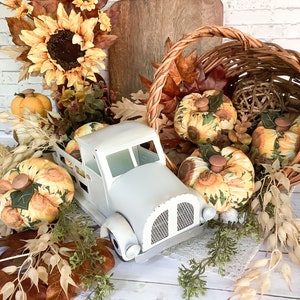 Puede incluir: Un cami&oacute;n de juguete de metal blanco que transporta calabazas de tela con patrones de girasoles, rodeado de hojas de oto&ntilde;o artificiales, girasoles, una cesta de mimbre tejida y una tabla de cortar de madera como decoraci&oacute;n oto&ntilde;al.