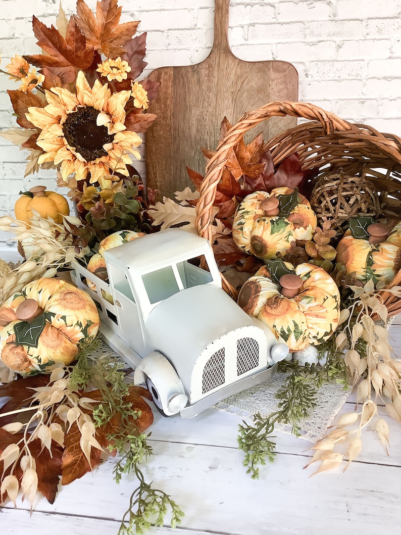 Puede incluir: Un cami&oacute;n de juguete de metal blanco que transporta calabazas de tela con patrones de girasoles, rodeado de hojas de oto&ntilde;o artificiales, girasoles, una cesta de mimbre tejida y una tabla de cortar de madera como decoraci&oacute;n oto&ntilde;al.