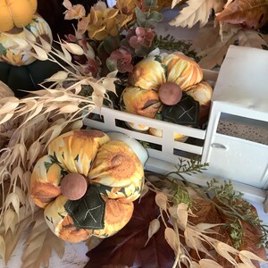 Puede incluir: Calabazas de tela con estampado de girasoles amarillos, centros de madera marr&oacute;n y hojas de tela verde oscuro. Las calabazas se exhiben junto a un modelo de cami&oacute;n de metal blanco, tallos de trigo seco y follaje de oto&ntilde;o artificial.