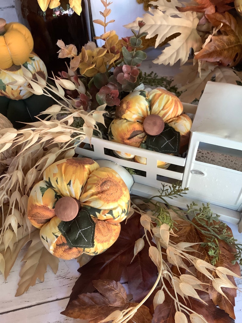 Puede incluir: Calabazas de tela con estampado de girasoles amarillos, centros de madera marr&oacute;n y hojas de tela verde oscuro. Las calabazas se exhiben junto a un modelo de cami&oacute;n de metal blanco, tallos de trigo seco y follaje de oto&ntilde;o artificial.