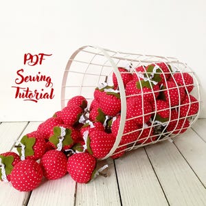 Può includere: Un cesto di filo bianco traboccante di fragole di stoffa rosse con pois bianchi. Le fragole cadono dal cesto su una superficie di legno bianco. Il testo "PDF Sewing Tutorial" è scritto in rosso su sfondo bianco.
