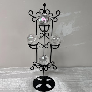 May include: A black metal crystal ball holder with four clear crystal spheres. The holder features an ornate design with curved accents and a circular base. The spheres are displayed at different levels, creating a visually appealing arrangement.