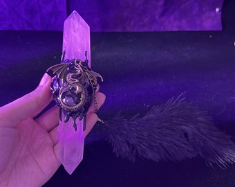 May include: A clear quartz crystal point with a black resin dragon pendant attached. The pendant has a bronze finish and a small amethyst stone in the center. A black feather is in the background.