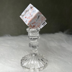 Puede incluir: Un candelabro de cristal transparente con base y tallo decorativos sostiene un cristal en forma de cubo. El cristal es blanco con patrones rojos y grises. Una pequeña etiqueta blanca con la letra "B" está adherida al cristal.