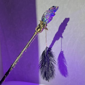 Op de afbeelding: Een zwarte veer hangt aan een gouden ketting die is bevestigd aan een toverstaf met een drakenkop en een regenboogkleurige kristallen top.