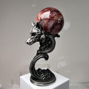 May include: A decorative sculpture featuring a detailed silver wolf head supporting a red and brown crystal sphere. The sculpture has an ornate, curved base and is displayed on a white pedestal. The crystal sphere is approximately 3 inches in diameter.