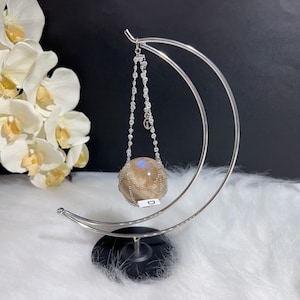 May include: A decorative sphere, suspended by a silver chain from a crescent-shaped stand. The sphere has a light brown color with a blue glow. The stand is silver and sits on a black base. White flowers are in the background.