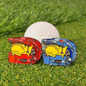 Può includere: Due segnaposto per palline da golf a forma di casco, uno rosso e uno blu, con visiere gialle e un dettaglio bandiera da golf. Una pallina da golf bianca è dietro di loro su erba sintetica verde.