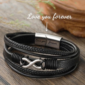 Könnte beinhalten: Schwarzes Lederarmband mit mehreren Strängen und einem silbernen Unendlichkeits-Symbol-Anhänger. Das Armband hat einen Magnetverschluss mit der Gravur "Love you forever".