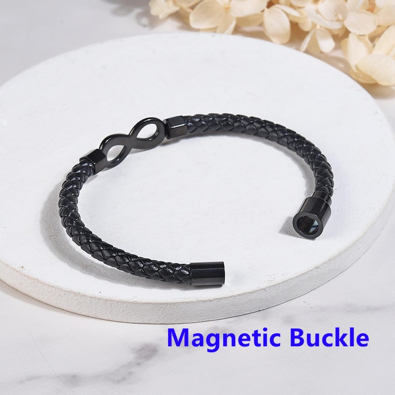 May include: Black leather bracelet with an infinity symbol charm and a magnetic buckle. The bracelet is on a white surface.