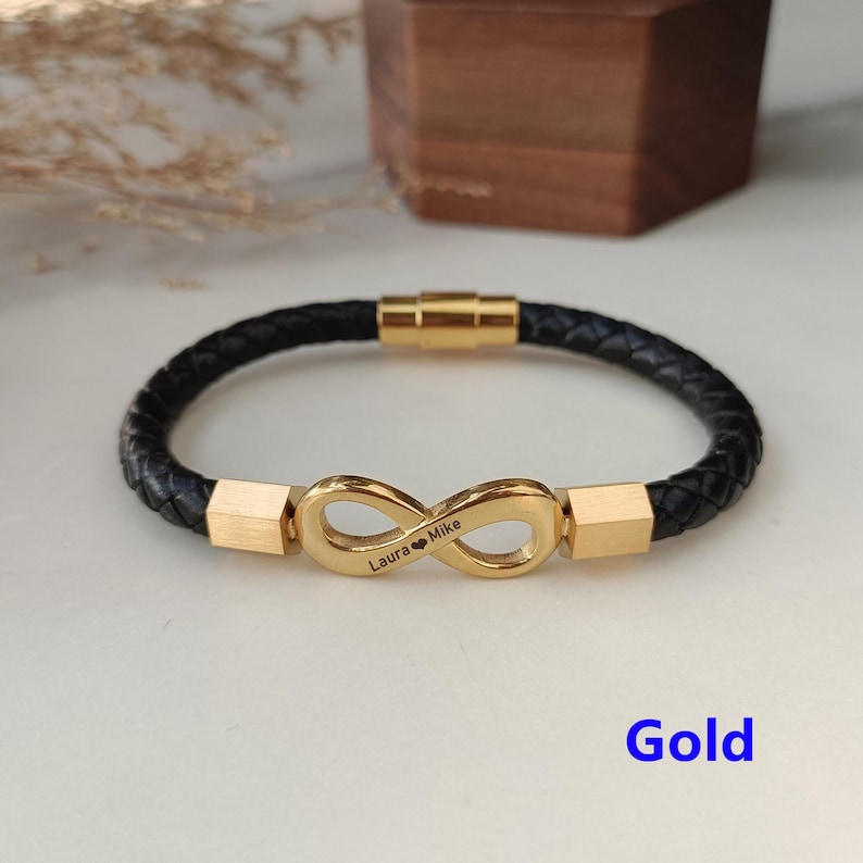 May include: A black leather bracelet with a gold infinity symbol charm. The charm is engraved with the names "Laura" and "Mike" and a heart symbol. The bracelet has a magnetic clasp.