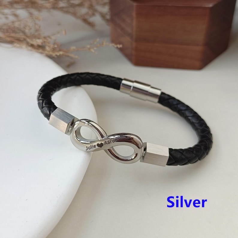 May include: A black leather bracelet with a silver infinity symbol charm. The charm is engraved with the names "Julie" and "Aaron" and a heart symbol.