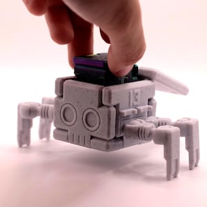 Könnte beinhalten: Ein hellgraues, 3D-gedrucktes Roboterspielzeug mit einem blockartigen Körper, kreisförmigen augenähnlichen Merkmalen und beweglichen Beinen. Eine Hand platziert eine schwarze und violette Komponente oben. Der Roboter hat die Nummer "13".