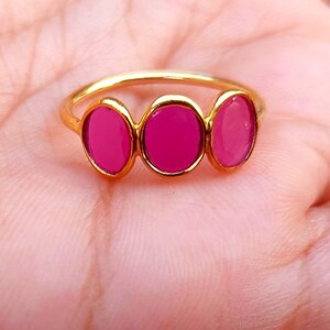 May include: A gold-toned ring featuring three oval-shaped, vibrant pink stones set horizontally in a row. The band is thin and metallic, holding the three matching pink cabochon-style stones securely in place across the top of the ring design.