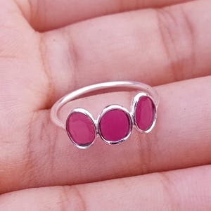 Könnte beinhalten: Ein silberner Ring mit drei kleinen, ovalen, magentafarbenen Einlagen, die horizontal entlang der Ringschiene angeordnet sind. Das Metallband ist glatt und poliert und bildet einen klaren Kontrast zu den leuchtend farbigen, runden Fassungen.