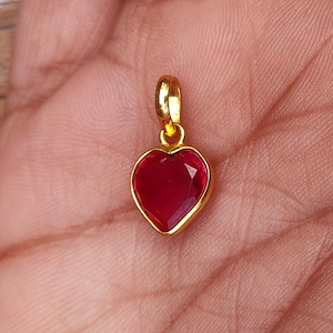 Könnte beinhalten: Ein goldfarbener herzförmiger Anhänger mit einem facettierten, tiefroten Edelstein. Der Anhänger ist an einer kleinen Schlaufe befestigt, die an einer Halskette oder einem Armband befestigt werden kann. Die Herzform ist ein klassisches Symbol der Liebe.
