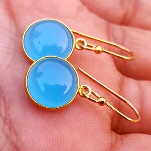Könnte beinhalten: Ein Paar runde, hellblaue Cabochon-Ohrringe in goldfarbenen Metallfassungen. Jeder Ohrring hat eine glatte, polierte Oberfläche und ist an einem einfachen goldfarbenen Haken zur Befestigung angebracht, was eine helle und gleichmäßige Farbe zeigt.