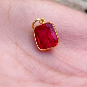 Könnte beinhalten: Ein rechteckiger Anhänger mit einem leuchtend roten facettierten Edelstein, der in einen polierten goldfarbenen Metallrahmen gefasst ist. Der Anhänger verfügt über einen kleinen runden goldfarbenen Verbindungsring an der Oberseite zur Befestigung.