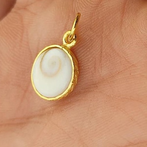 K&ouml;nnte beinhalten: Ein goldfarbener Anh&auml;nger mit einem ovalen, cremefarbenen Muscheldesign. Der Anh&auml;nger hat einen strukturierten Goldrand und eine kleine Schlaufe f&uuml;r eine Kette. Die Muschel hat ein Spiralmuster.